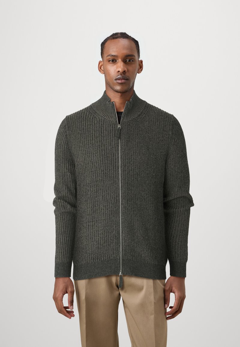 Dark green zip-up cardigan with ribbed texture, high collar, and long sleeves. Features silver zipper and finished hem. Worn over beige trousers.