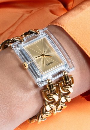 Orologio rettangolare trasparente con quadrante dorato, bracciale a maglie dorate e cassa trasparente, indossato su un polso con tessuto arancione sullo sfondo.