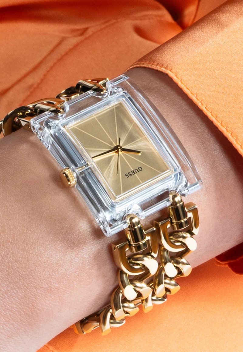 Klart rektangulært ur med guldskive, guld kædelænke og en gennemsigtig kasse, båret på et håndled med orange stof i baggrunden.