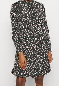 Robe noire à manches longues, motif floral en rose et blanc, devant boutonné, taille plissée et coupe décontractée. Texture de tissu lisse.