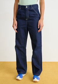 Mörkblå denim breda byxor med synliga vita sömmar, hög midja och framfickor, kombinerat med blå sneakers för träning.
