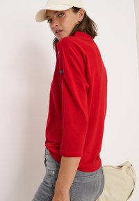 Haut rouge à manches longues avec un col montant, ornée de boutons noirs sur les épaules, associée à un jean gris et un sac de couleur claire.