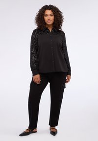 Camicia nera con colletto e maniche di paillettes, abbinata a pantaloni neri con dettagli texturizzati. Tessuto liscio con vestibilità morbida.