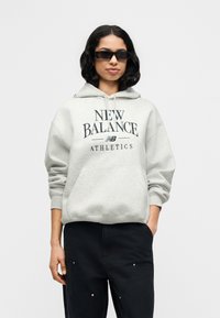 Ανοιχτόγριζη μπλούζα με κουκούλα και μπροστινή τσέπη καγκουρό, με μαύρο κείμενο "NEW BALANCE ATHLETICS". Συνδυάζεται με μαύρο παντελόνι και γυαλιά ηλίου.