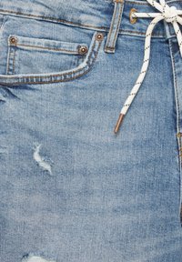 Vaaleansiniset farkkushortsit, joissa on kulutettuja yksityiskohtia, valkoisella nyörillä ja kuparinvärisillä varusteilla vyötäröllä ja taskuissa.