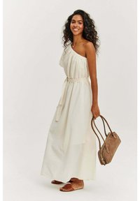 Maxi šaty z svetlej béžovej látky s dizajnom jedného ramena, všitou pásovou časťou a voľnou siluetou. Skombinované s hnedou kabelkou.