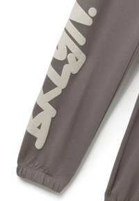 Graue Jogginghosen mit elastischen Bündchen, mit großem weißem Grafiktext entlang des linken Beins. Hergestellt aus weichem, dehnbarem Stoff.