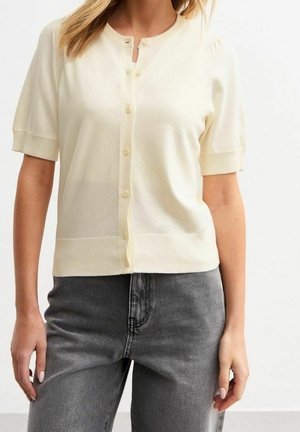 Femme portant un cardigan crème à manches courtes boutonné, associé à un jean noir délavé taille haute.