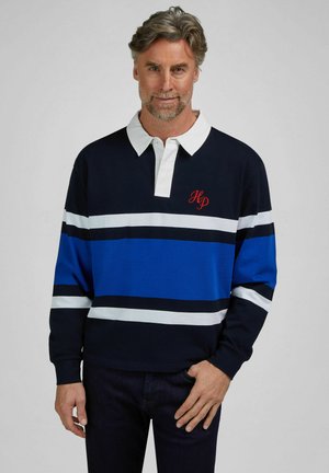 Mann mit grauen Haaren, der ein marineblau, blau und weiß gestreiftes Rugbyshirt mit weißem Kragen und rotem "HP"-Monogramm auf der Brust trägt.