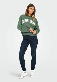 Groene sweatshirt met "NUMBER NINE" in het wit gedrukt, gecombineerd met donkere skinny jeans en witte high-top sneakers. Casual pasvorm en stijl.