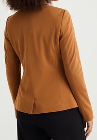 Brun skräddarsydd kavaj med en figurnära design, strukturerade axlar och en subtil textur. Har en slits bak och långa ärmar.