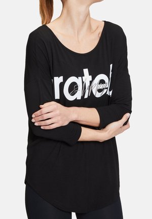 Femme croisant les bras, portant une chemise noire à manches longues avec un grand texte blanc partiellement lisible "grateful" sur le devant.