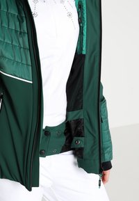 Veste isolante verte avec une fermeture éclair à l'avant, des boutons-pression, des poches latérales, des textures contrastantes et une doublure intérieure noire.