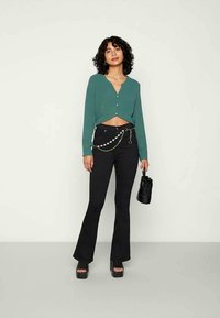 Top verde cropped de manga longa com botões, combinado com calças flare pretas. Inclui um cinto de corrente e uma mala preta, usado com sapatos de salto plataforma.