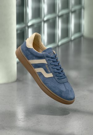 Blue suede sneaker with beige stripes, brown sole, and laces, floating indoors on a polished concrete floor near glass blocks.