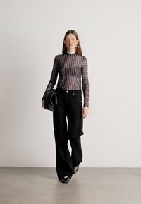 Top trasparente a righe nere a maniche lunghe abbinato a pantaloni neri larghi a vita alta. La modella tiene una morbida borsa nera; ambiente minimalista.
