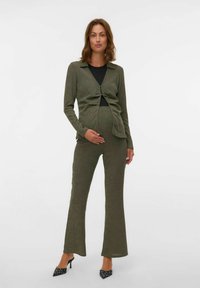 Zwanger vrouw in olijfgroene ribgebreide cardigan en broek, met een zwarte top eronder en zwarte pumps met stippen, staande tegen een witte achtergrond.