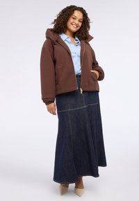 Jachetă cu fermoar, cu glugă, de culoare maro, realizată dintr-un material moale, asortată cu o fustă lungă din denim albastru, cu detalii pe cusături. Purtată cu tocuri de un bej deschis.