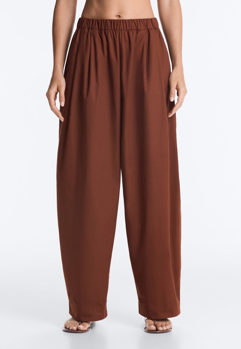 Personne portant un pantalon marron ample taille haute avec une ceinture élastique et des sandales à bouts ouverts sur un fond uni.