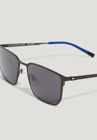 Occhiali da sole quadrati con lenti grigie, montatura sottile in metallo nero, estremità delle stanghette blu e piccolo logo Tommy Hilfiger sulle braccia.