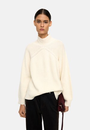 Biały sweter z dzianiny z wysokim kołnierzem i prążkowaną teksturą, w zestawie z ciemnymi spodniami i małą bordową torbą. Prosty design, bez wzorów.