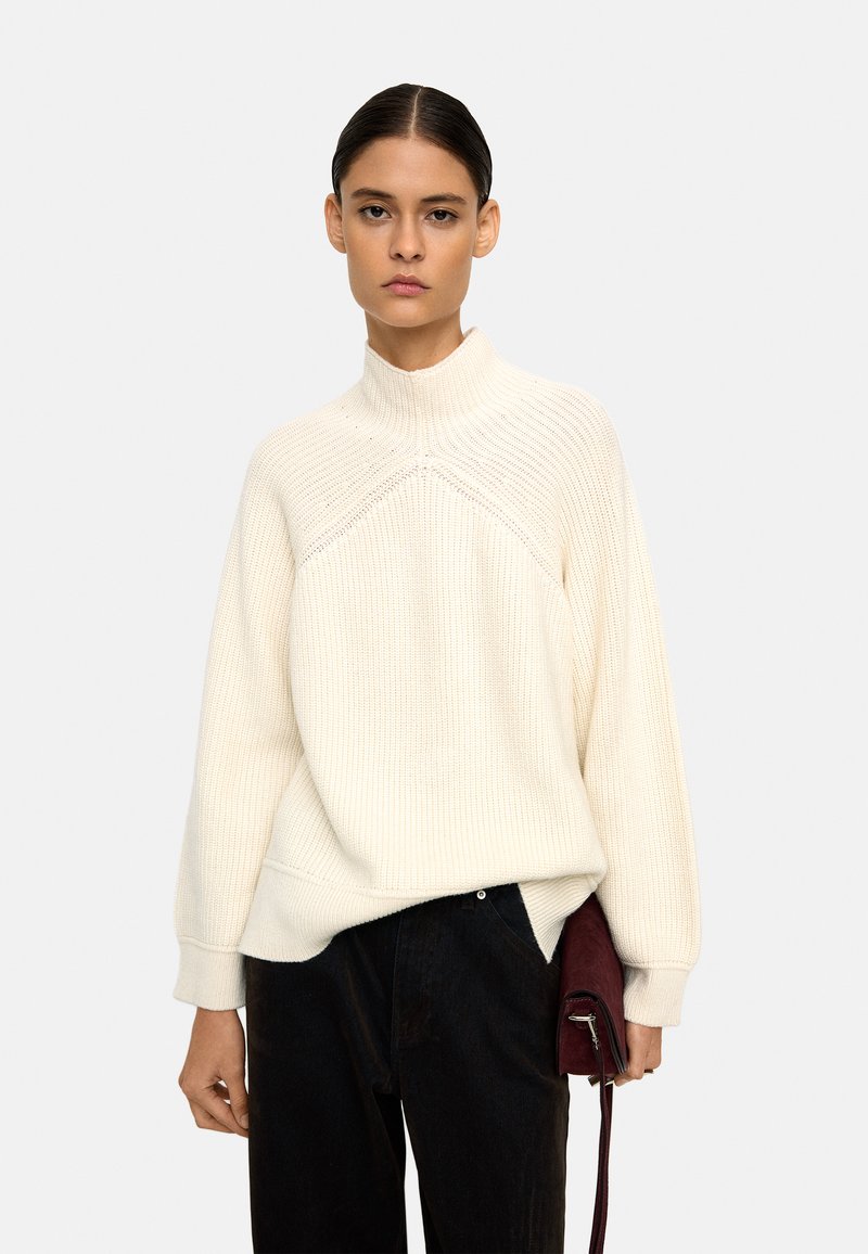 Biały sweter z dzianiny z wysokim kołnierzem i prążkowaną teksturą, w zestawie z ciemnymi spodniami i małą bordową torbą. Prosty design, bez wzorów.