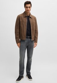 Chaqueta de ante marrón con cremallera frontal, camiseta negra, jeans grises y zapatillas negras. La chaqueta cuenta con un cuello y bolsillos laterales.