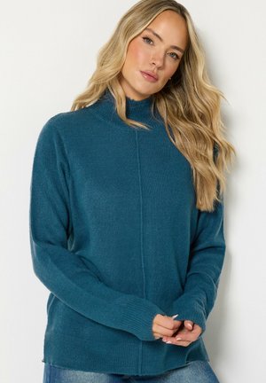 Femme aux longs cheveux blonds portant un pull en maille bleu sarcelle et un jean bleu, debout devant un fond blanc uni.