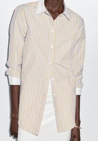 Yellow and white striped shirt with a button-down collar, long sleeves, and a curved hem. Fabric appears smooth with white accents at cuffs.