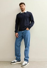 Marineblå långärmad polo med vit krage, matchad med ljusblå raka jeans och grå, vita och svarta sneakers mot en neutral bakgrund.