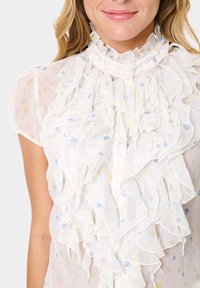 White ruffled blouse with floral patterns in blue and yellow, featuring a high collar and button closure, made from lightweight fabric.
