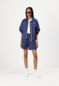 Maritime jas met gele accenten en een witte rits, gecombineerd met marineblauwe cargoshorts en witte sneakers. Model draagt rode zonnebril.