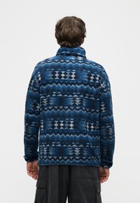 Άνδρας που φοράει μπλε μπουφάν από φλις με σχέδια και σκούρο παντελόνι τύπου cargo, εμφανίζεται από την πίσω πλευρά μπροστά σε απλό λευκό φόντο.