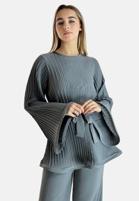 Top gris plisado con mangas acampanadas, tejido texturizado, cuello redondo y un lazo a juego en la cintura. Combinado con pantalones grises.