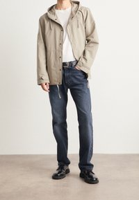 Osoba ubrana w beżową kurtkę z kapturem, białą koszulę, ciemnoniebieskie dżinsy, czarny pasek i czarne, wypastowane buty stoi na szarym podłodze.