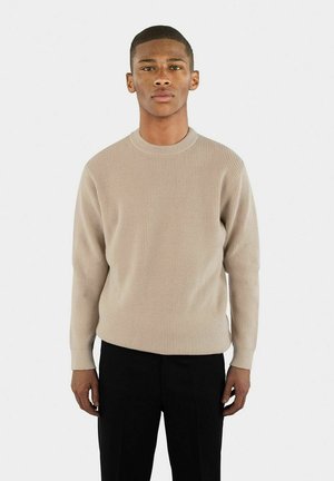 Beige ribbat stickad tröja med rund halsringning, långa ärmar och avslappnad passform, tillsammans med svarta byxor.