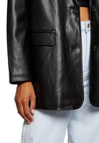 Käsi lepoaa musta nahkableiseri taskun vieressä, joka on päällä vaaleansiniset korkeavyötäröiset farkut, paljastaen vatsan.