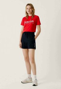 Rode t-shirt met "Red Bull Racing" logo, zwarte shorts met elastische tailleband, gecombineerd met witte sneakers en sokken, staand op een neutrale achtergrond.