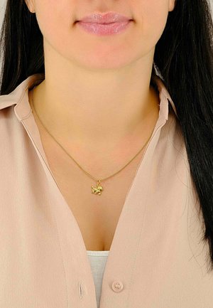 Collier en pendentif en or avec un charme en forme de chien sur une fine chaîne, porté sur un chemisier beige clair et un haut blanc. Texture lisse, éclat subtil.