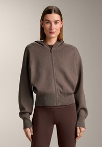 Bruine capuchontrui met ritssluiting, verzonken schouders, geribde manchetten en zoom, gemaakt van een zachte stof. Draagt het over een donkerbruine top en legging.