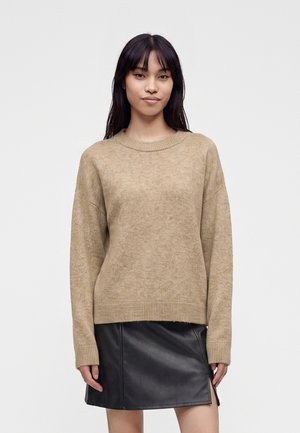 Vrouw die een beige gebreide trui en een zwarte leren rok draagt, staand tegen een effen lichte achtergrond.