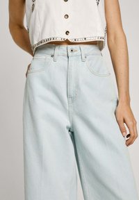 Vaaleansiniset korkeavyötäröiset väljät farkut, joissa on hienovaraiset ompeluyksityiskohdat ja etutaskut. Yhdistettynä lyhytvartiseen valkoiseen nappipaitaan.