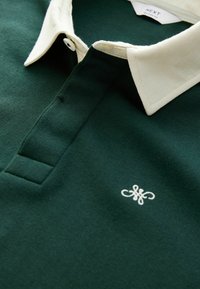 Tamnozelena polo majica s kontrastnim kremastim ovratnikom, bijelim gumbom i izvezenim logotipom na prsima. Pamuk, glatka tekstura.