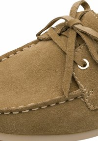 Chaussure de style mocassin en daim marron avec un bout rond, des coutures décoratives et des lacets passés dans un œillet de couleur or.