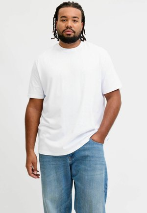 Mand med flettet hår iført en enkel hvid t-shirt og blå jeans, stående med den ene hånd i lommen foran en hvid baggrund.