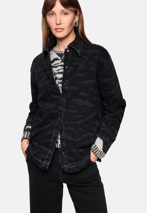 Black denim jacket with a subtle camouflage pattern, featuring a button closure, side pockets, and a relaxed fit over a patterned sweater.