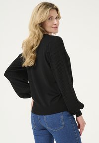 Schwarzer Pullover mit gerippten Texturärmeln und taillierter Passform. Paar blaue Jeans mit Rücktaschendetails. Neutraler Hintergrund.