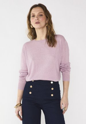 Donna con capelli castani mossi che indossa un maglione viola chiaro e pantaloni scuri a vita alta con bottoni dorati, in piedi con una mano lungo il corpo.