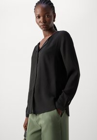Blusa nera a maniche lunghe con scollatura a V e chiusura a bottoni. Tessuto morbido; presenta una vestibilità rilassata e fessure laterali. Abbinata a pantaloni verdi.