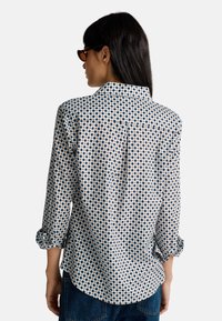 Una donna che indossa una camicia a maniche lunghe con un motivo e i polsini arrotolati, con occhiali da sole scuri, in piedi di spalle rispetto alla macchina fotografica.
